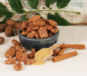 Cinnamon and sugar coated pecans in a bowl, surrounded by shelled pecans, unshelled pecans, and cinnamon sticks against a wooden background.