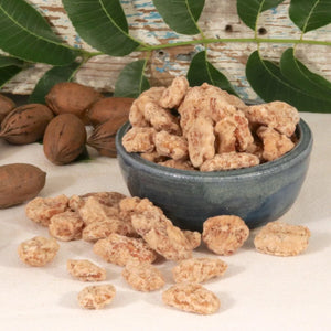 A bowl of praline pecans surrounded by unshelled pecans, and shelled praline pecans against a wooden background.