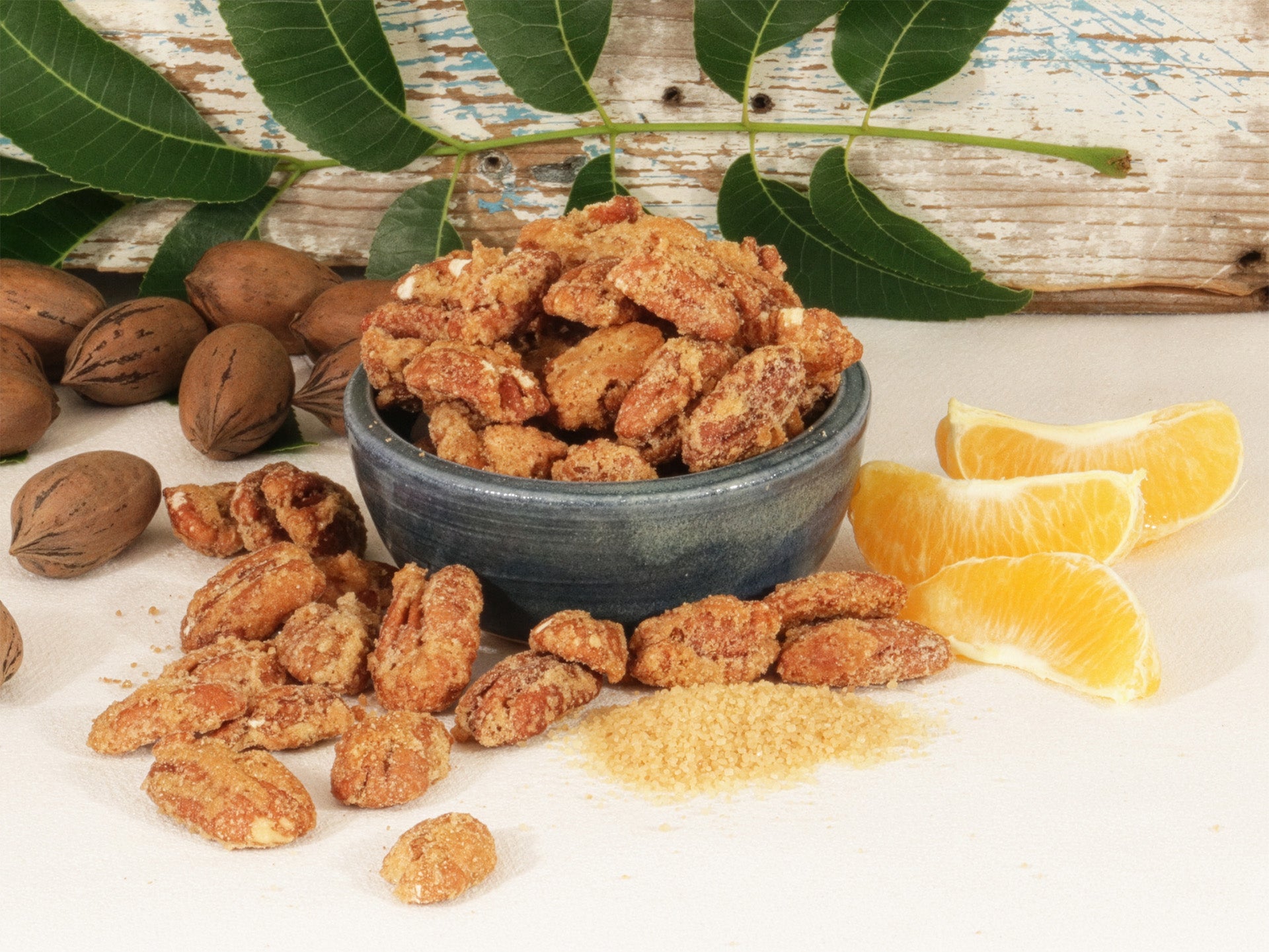 Shelled pecans with a sweet orange seasoning in a blue bowl, surrounded by unshelled pecans, loose shelled pecans with the sweet orange seasoning, and loose orange slices against a wooden background.