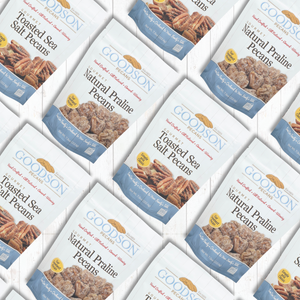 Multiple bags of Goodson Natural Praline Pecans arranged in a repeating diagonal pattern on a white background. The bags are white with blue bands, featuring a photo of toasted sea salt pecans and a yellow circular badge.